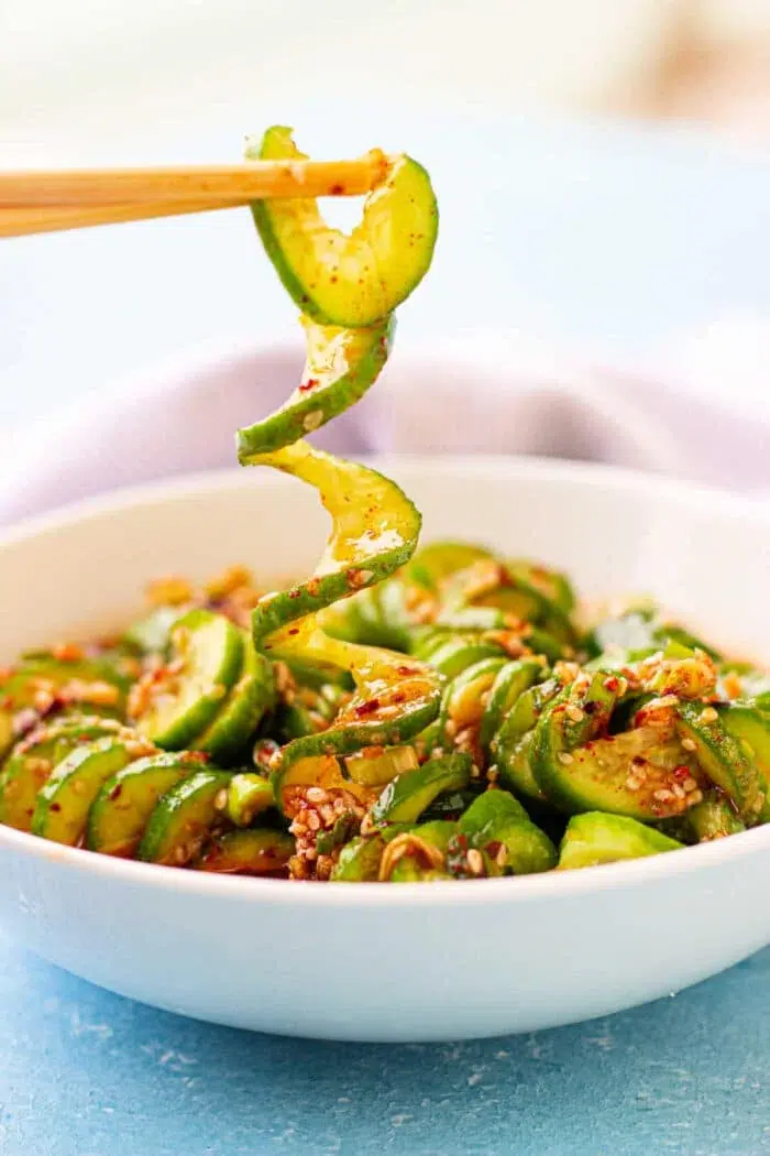 Cucumber ribbon salad in a white bowl with sesame seeds and chili flakes, held by chopsticks.