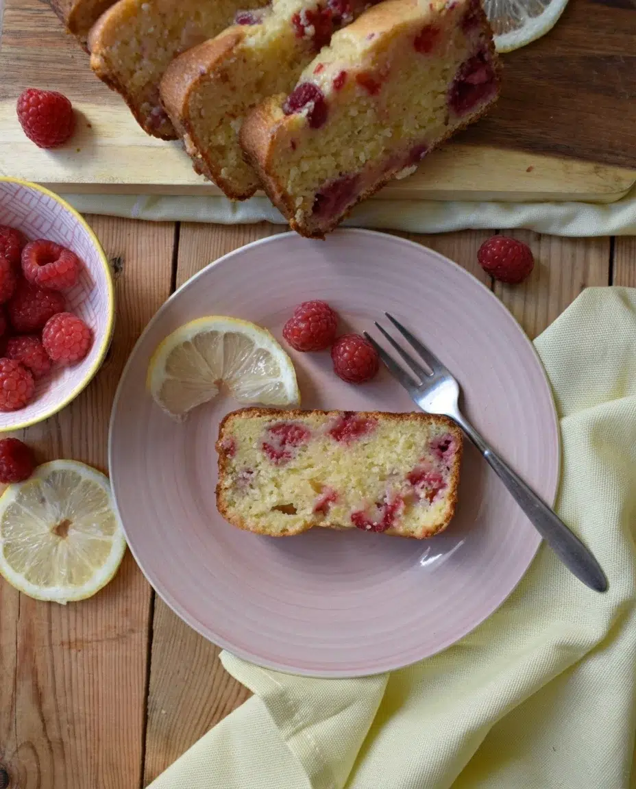 RASPBERRY-LEMON-LOAF-CAKE-IMAGE10-683x1024.jpg Slice of raspberry-lemon loaf cake on a pink plate with lemon wedges and raspberries.