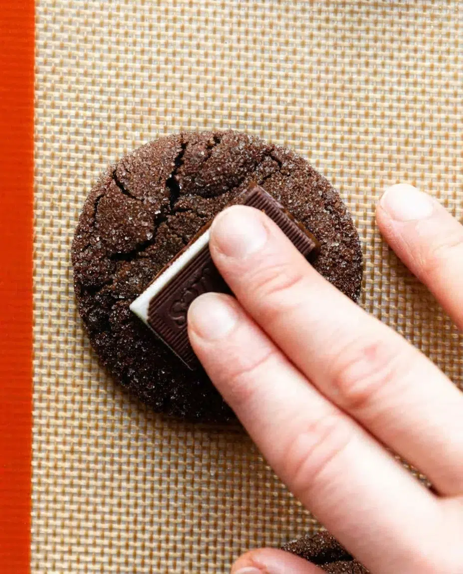 pressing-swirling-andes-mint-on-cookies.jpg Close-up of a dark chocolate cookie being topped with a rectangular chocolate bar on a woven placemat.