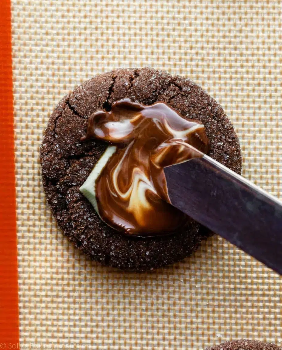 pressing-swirling-andes-mint-on-cookies-2.jpg close-up of a dark chocolate cookie with glossy melted chocolate being spread by a knife on a woven placemat