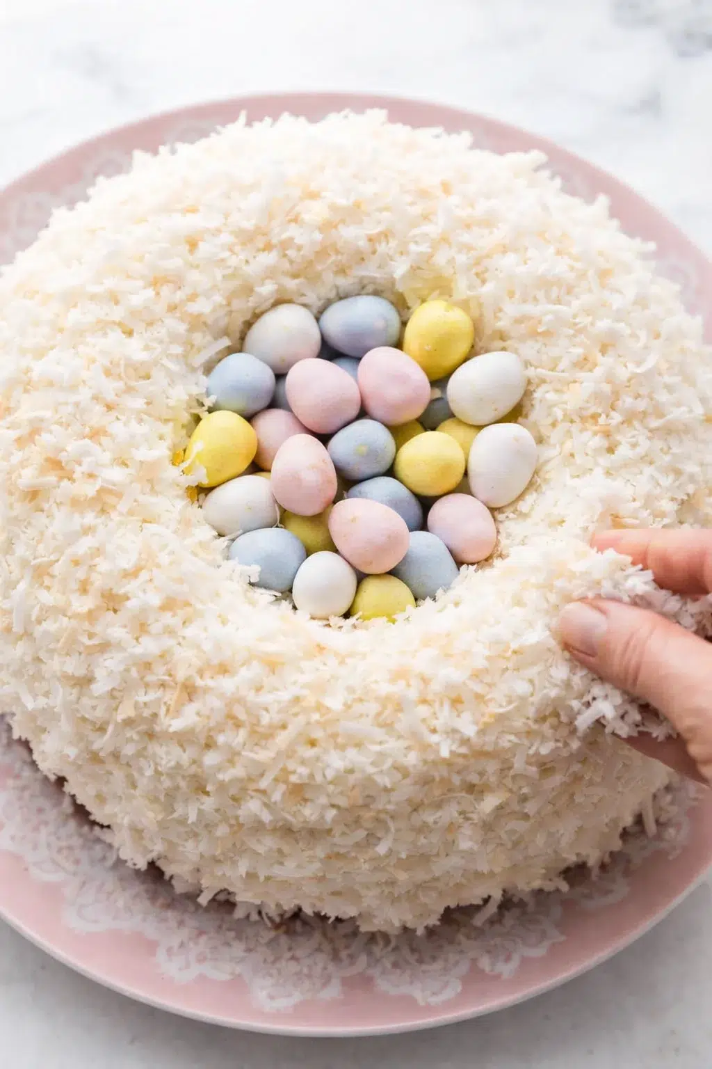 Hands pressing toasted shredded coconut onto frosted cake while center filled with candy eggs.