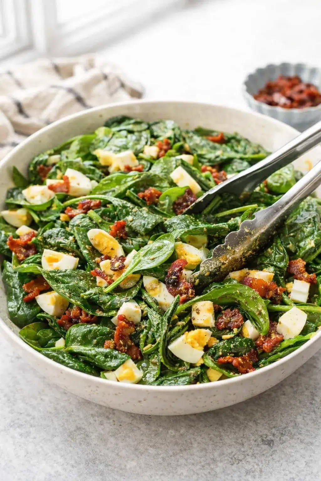 Large bowl of baby spinach mixed with crumbled bacon and chopped eggs, dressing being drizzled