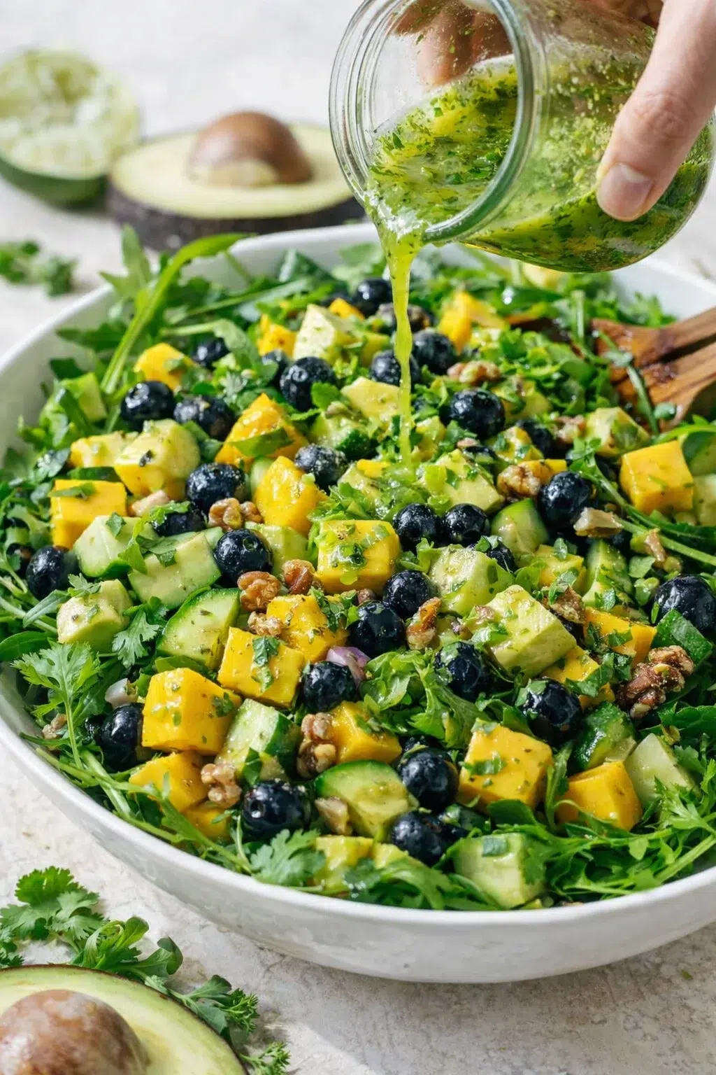Sliced avocado and blueberries being added, vinaigrette drizzled over salad while hands toss gently