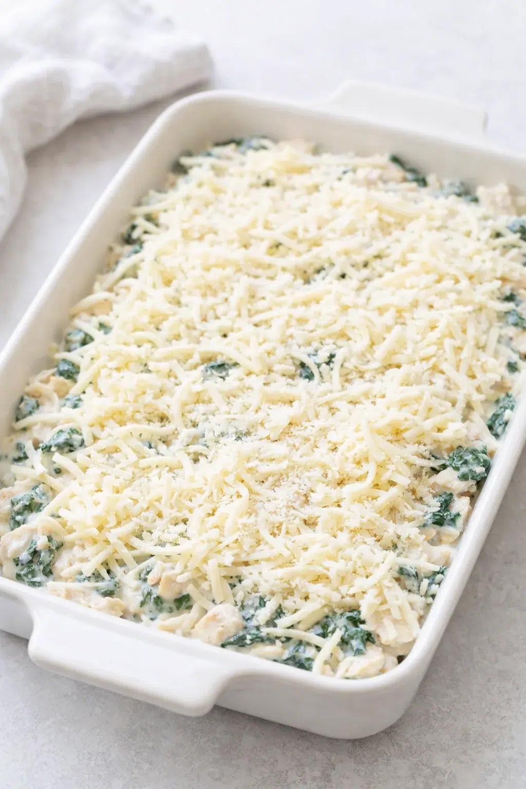 Casserole dish filled with chicken-spinach mixture, hands smoothing mixture and sprinkling cheeses