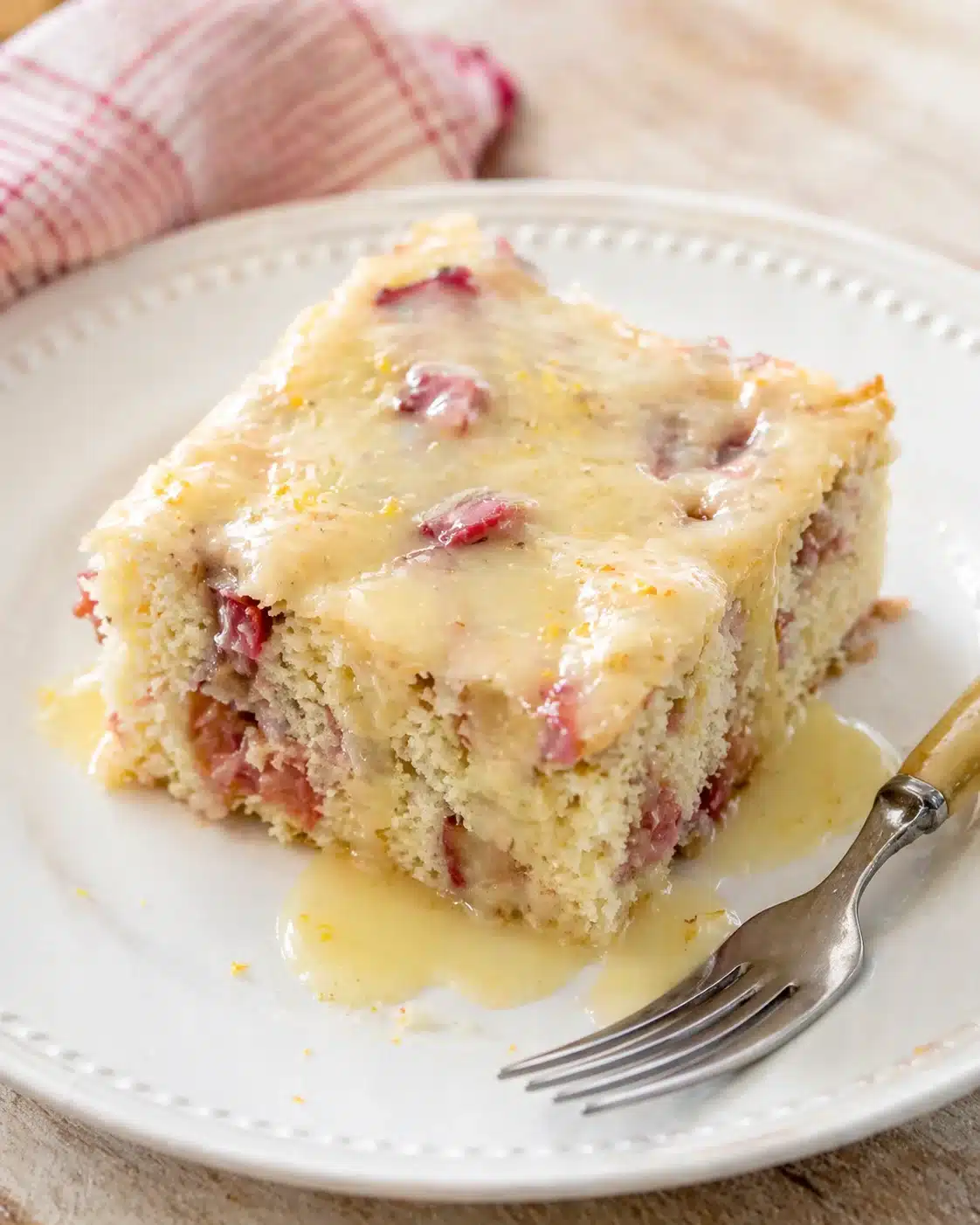 A slice of light-yellow cake with red fruit chunks, topped with pale glaze on a white plate.