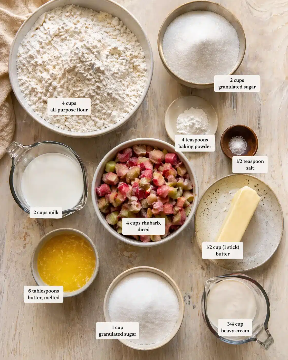 Overhead view of baking ingredients arranged on a wooden surface: flour, rhubarb, sugar, butter, milk, and cream.