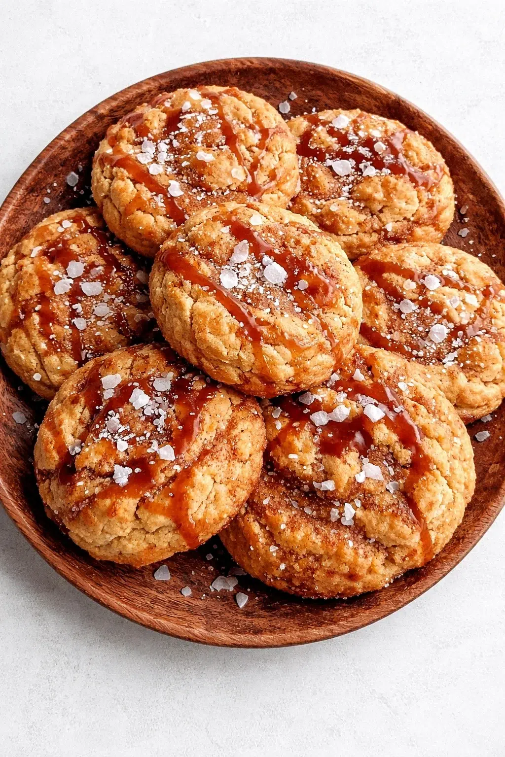 A wooden plate piled with caramel-drizzled, salted cookies on a pale background.