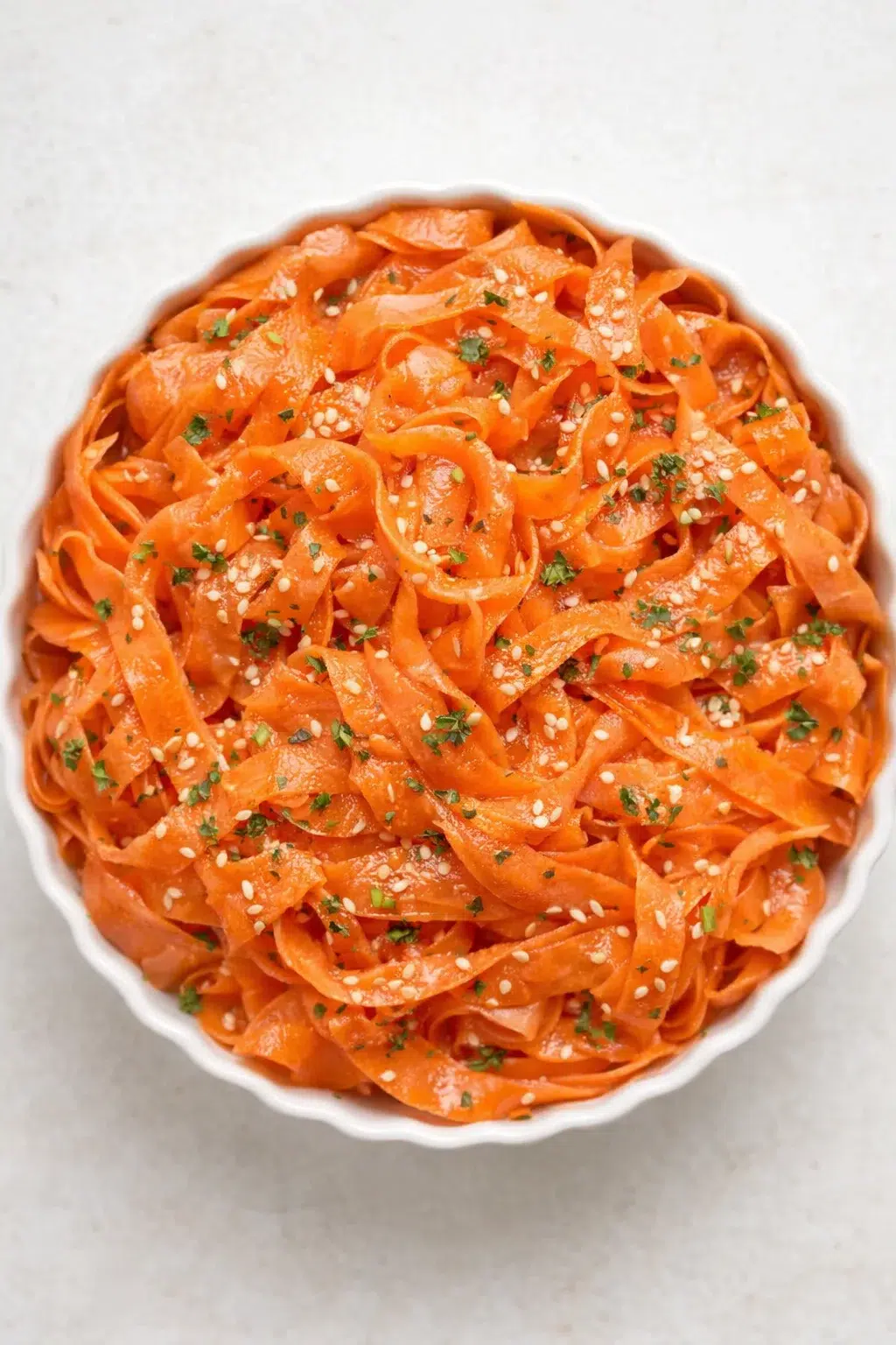 Top-down view of orange carrot ribbons in a white scalloped bowl, sesame seeds and chopped herbs.