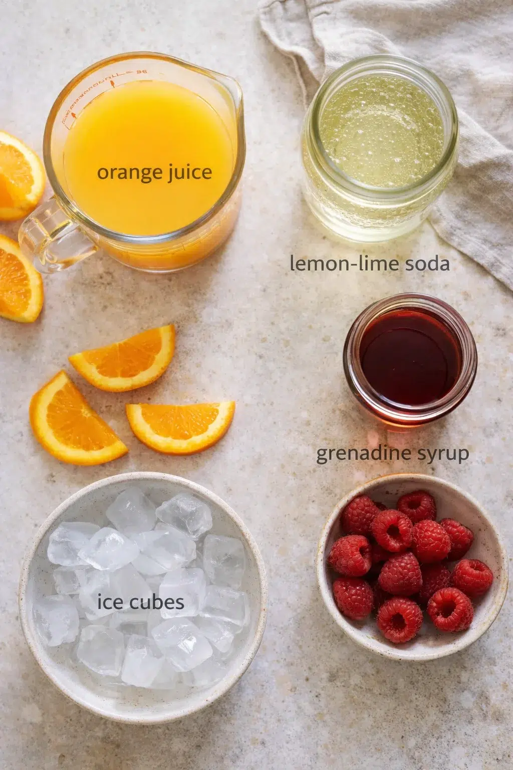Overhead view of orange juice, lemon-lime soda, grenadine syrup, ice cubes, raspberries, and orange slices arranged on a countertop.