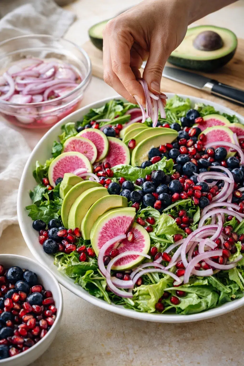 Vibrant salad bowl with greens, avocado slices, watermelon radish, blueberries, pomegranate seeds, and red onion.