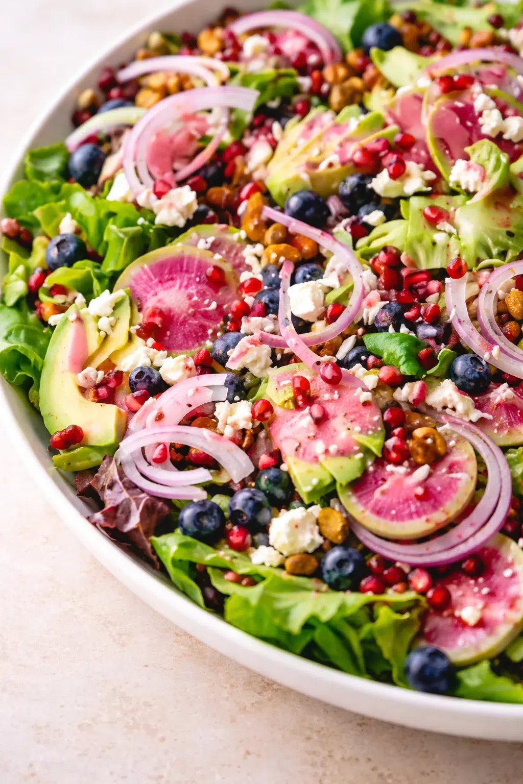 vibrant salad in a white bowl with greens, avocado, watermelon radish, blueberries, feta, pomegranate seeds, and red onion