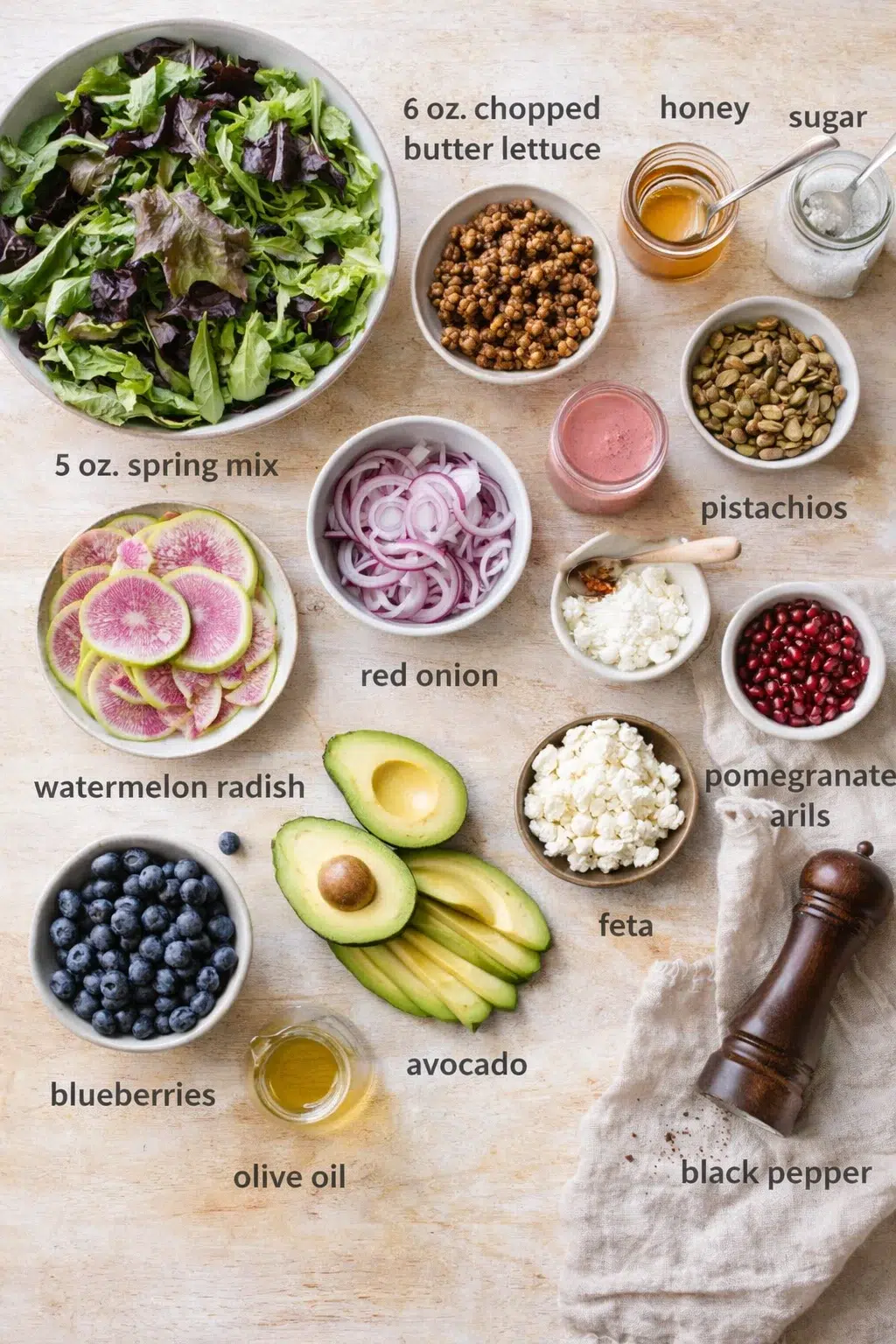 Overhead shot of colorful salad ingredients labeled in bowls on a light wood surface.