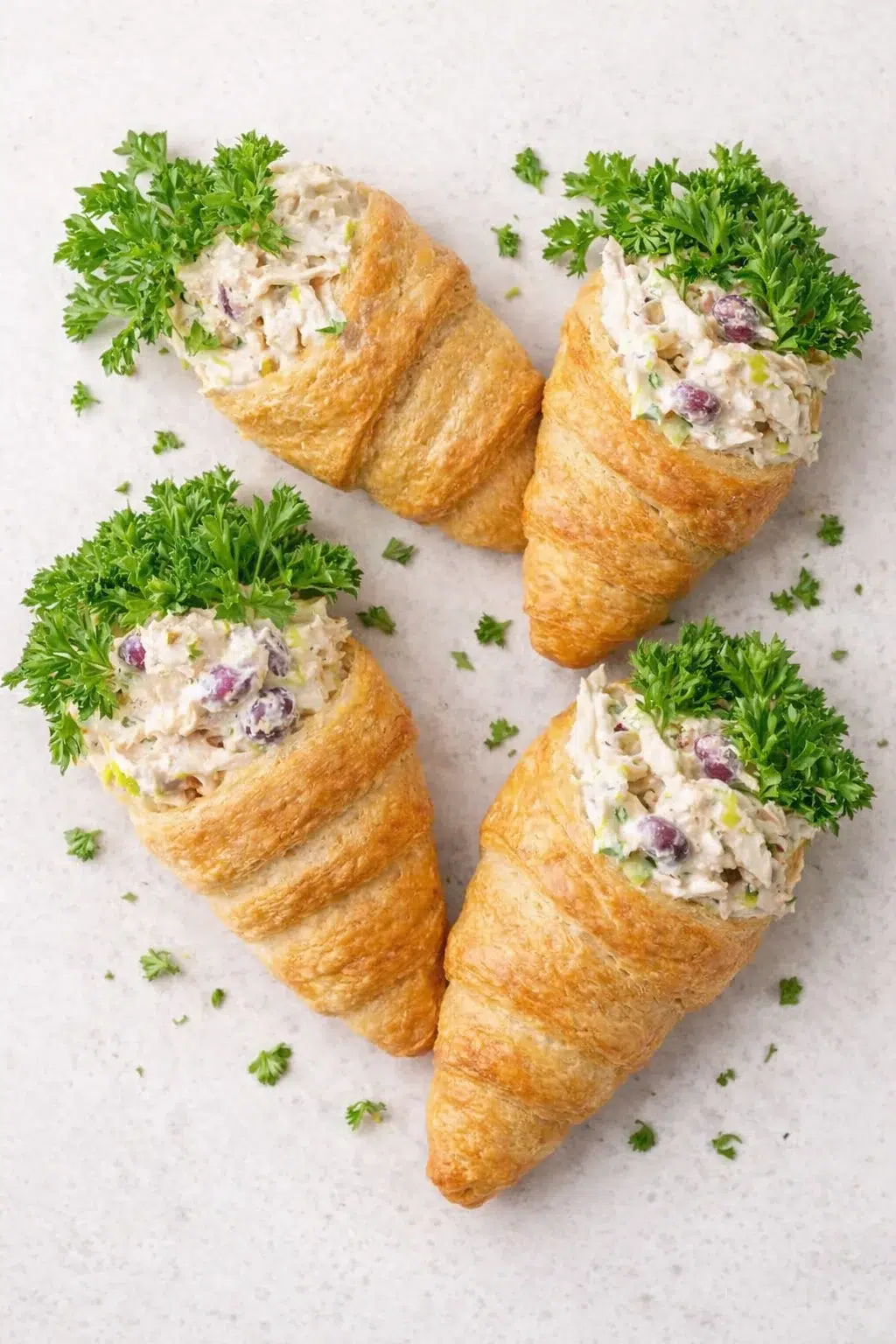 Four croissant shells filled with creamy chicken salad, topped with parsley on a light gray surface.