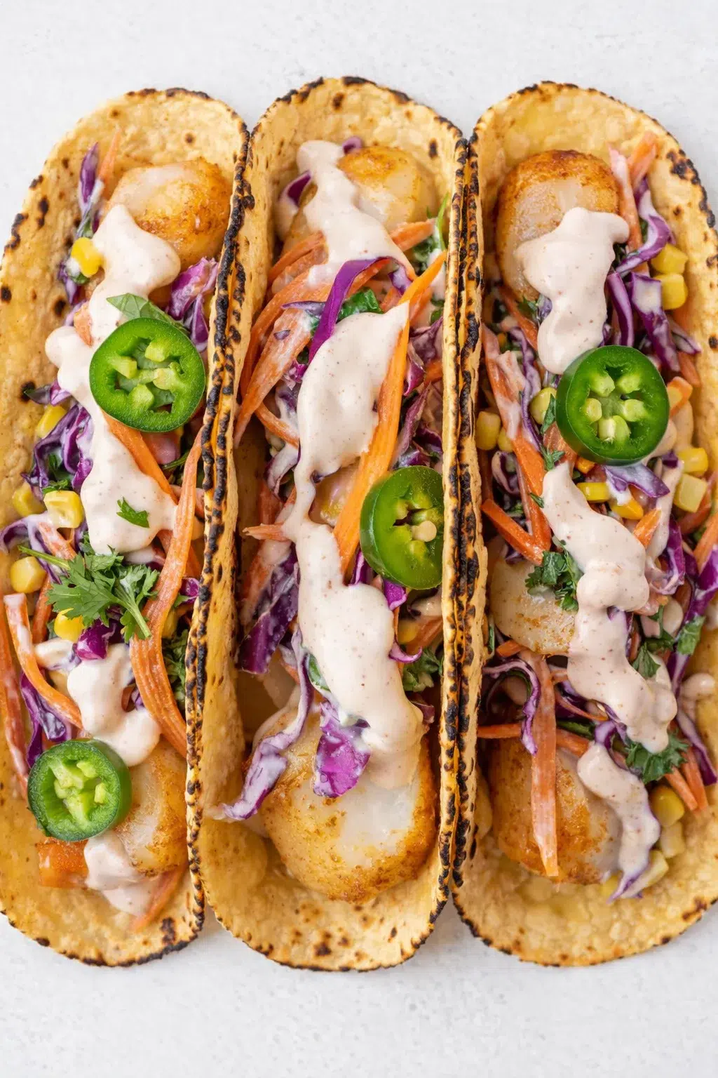 three crispy fish tacos with colorful slaw, jalapeño slices, and creamy sauce on a light background.