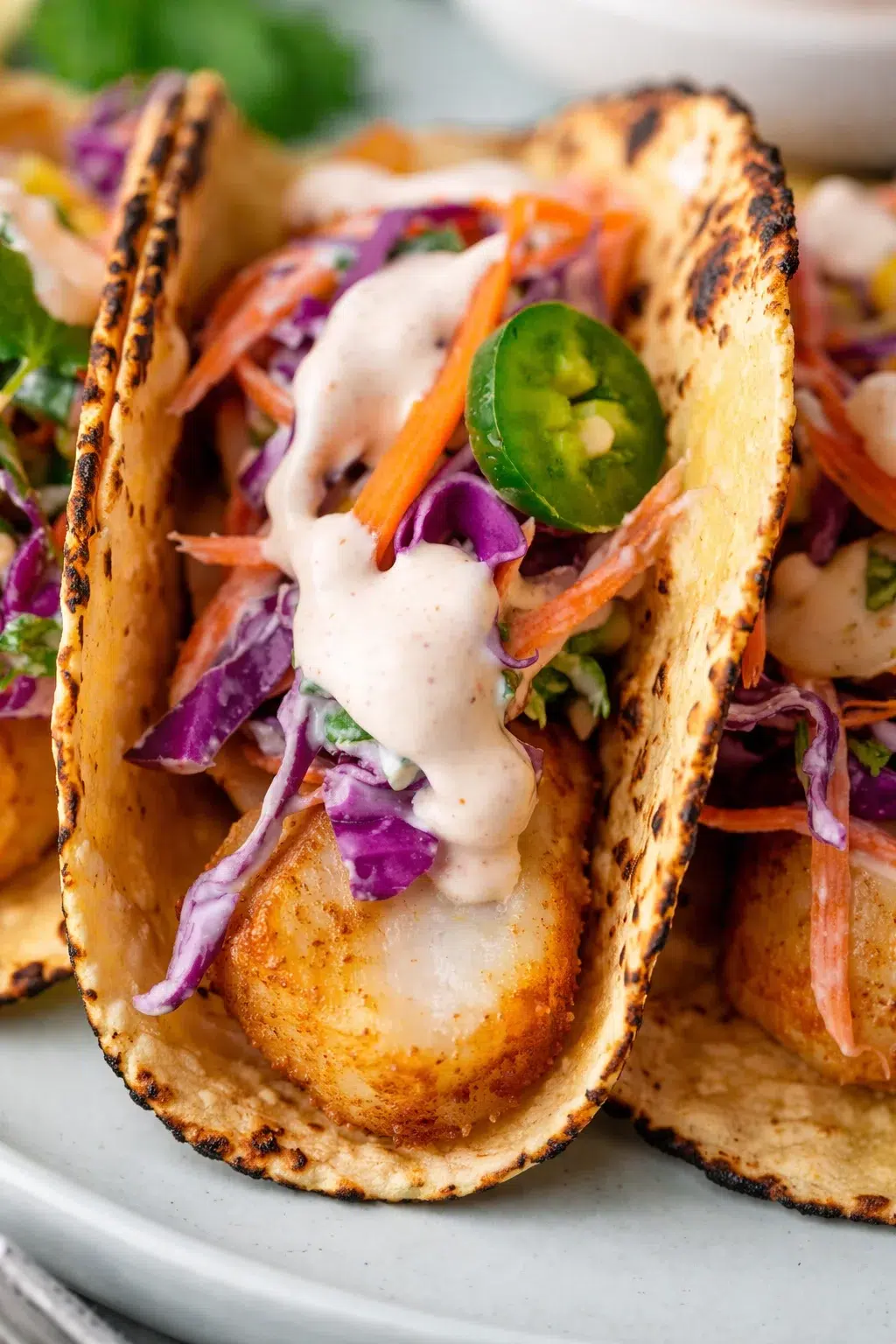 Close-up of two crispy fish tacos with purple cabbage slaw, carrot, jalapeño, and creamy sauce.