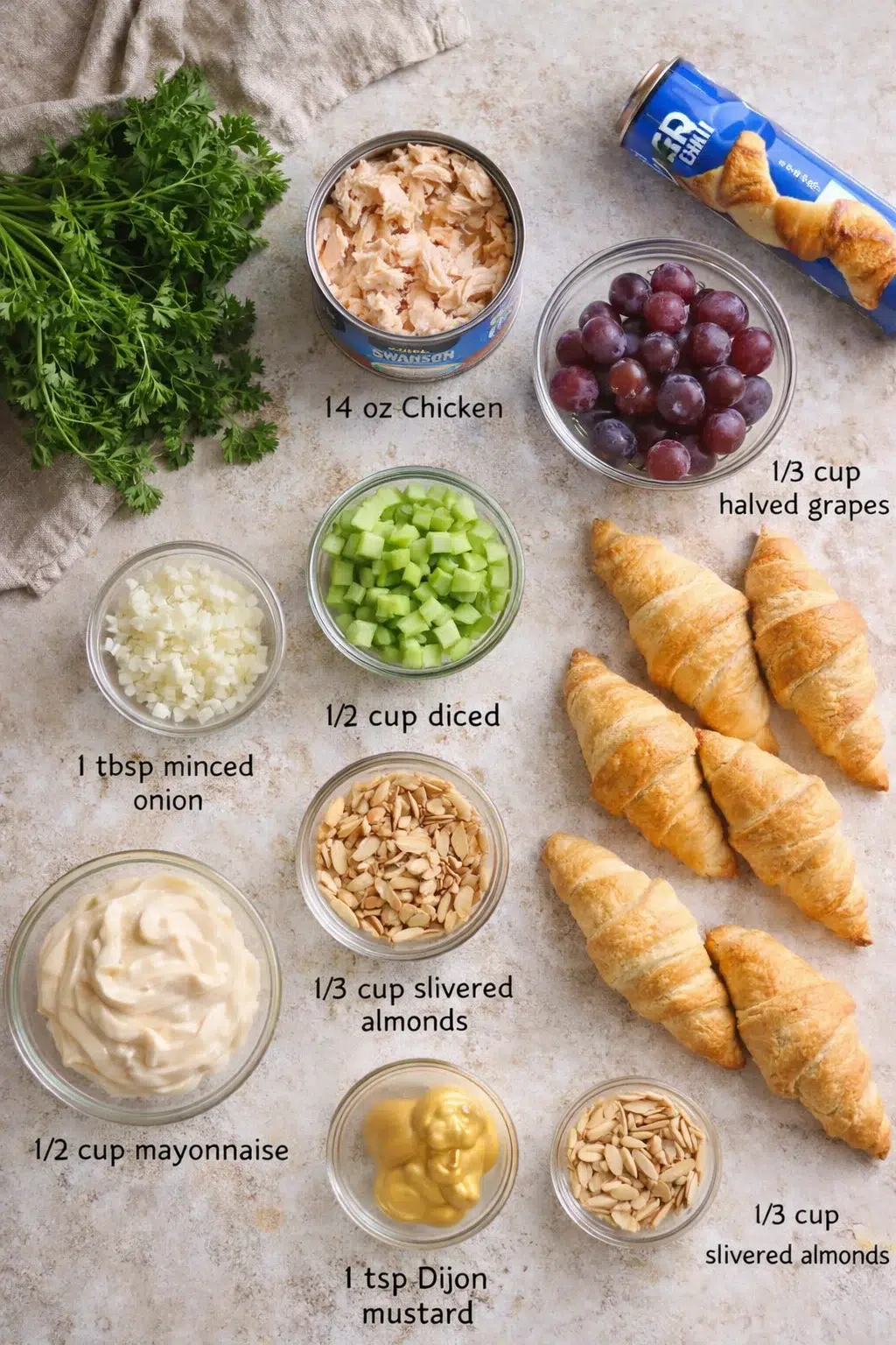 Top-down view of parsley, tuna can, halved grapes, croissants, and small bowls of chopped onion, cucumber, almonds, mayo, and mustard.