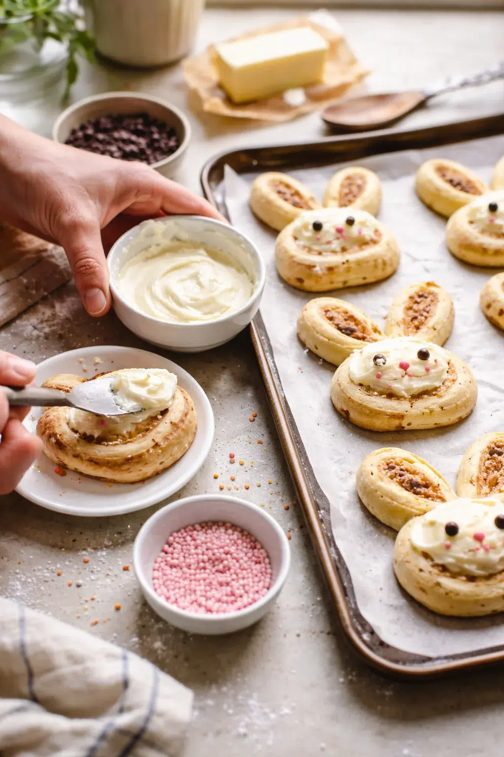 A hand spreads creamy frosting onto bunny-shaped pastries on a parchment-lined baking sheet.
