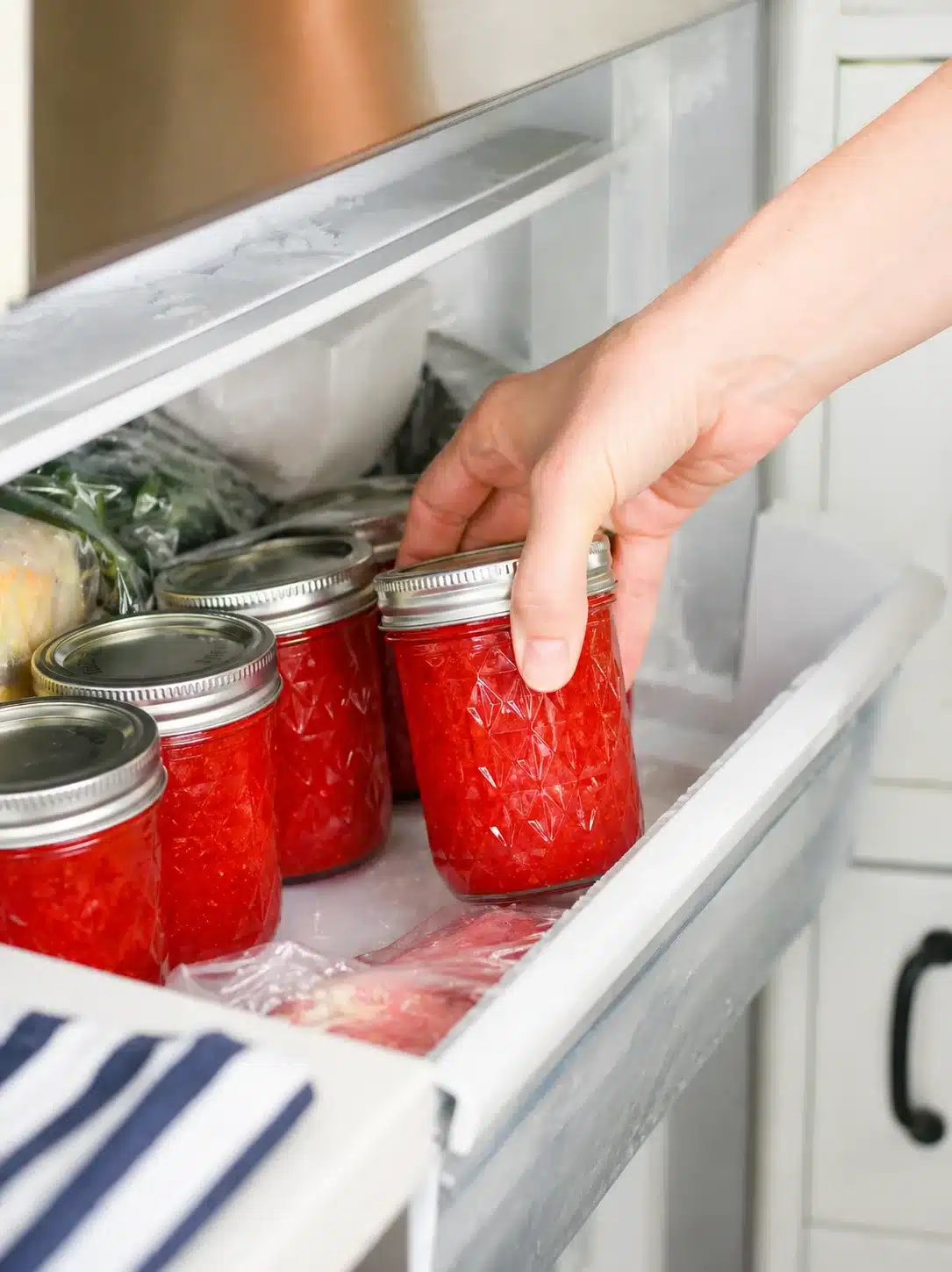 hand reaches to place bright red tomato sauce jars into a frosty freezer shelf