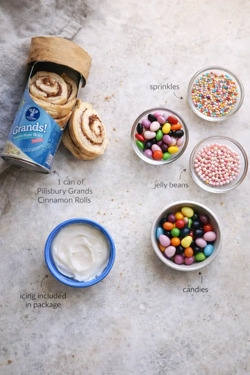 Overhead view of cinnamon rolls in a can with icing, bowls of jelly beans, sprinkles, and candies.
