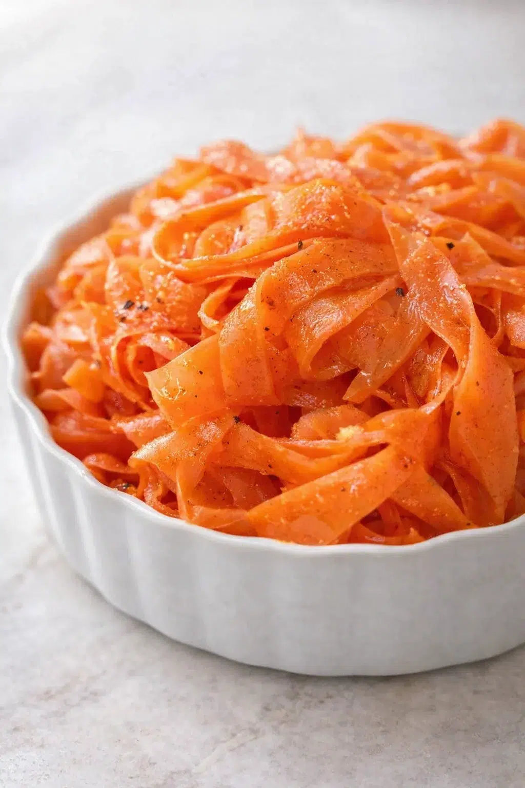 Bright orange carrot pasta ribbons in a white scalloped bowl, glossy with a light oil