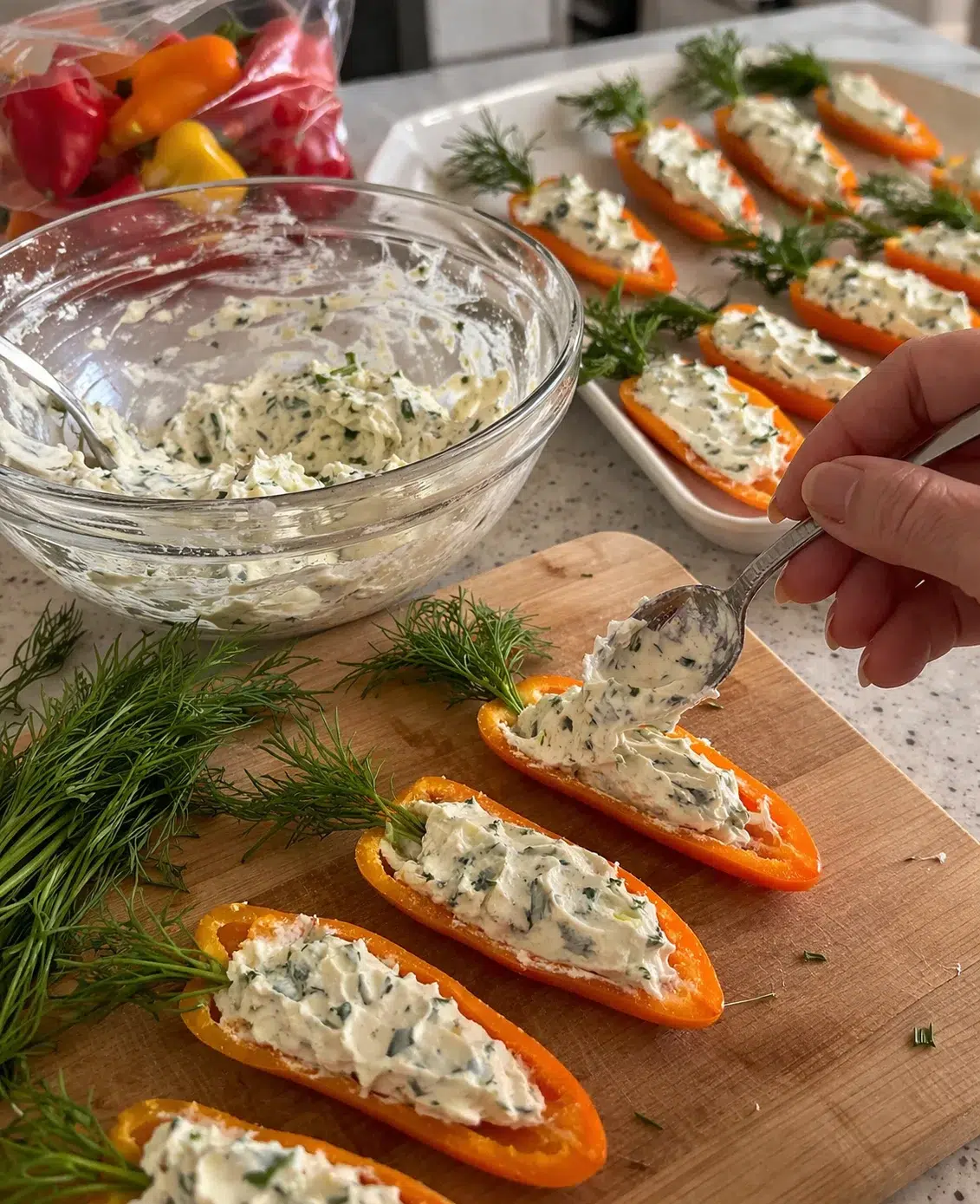 hand scooping herb-filled cream cheese onto orange pepper halves on a wooden cutting board.