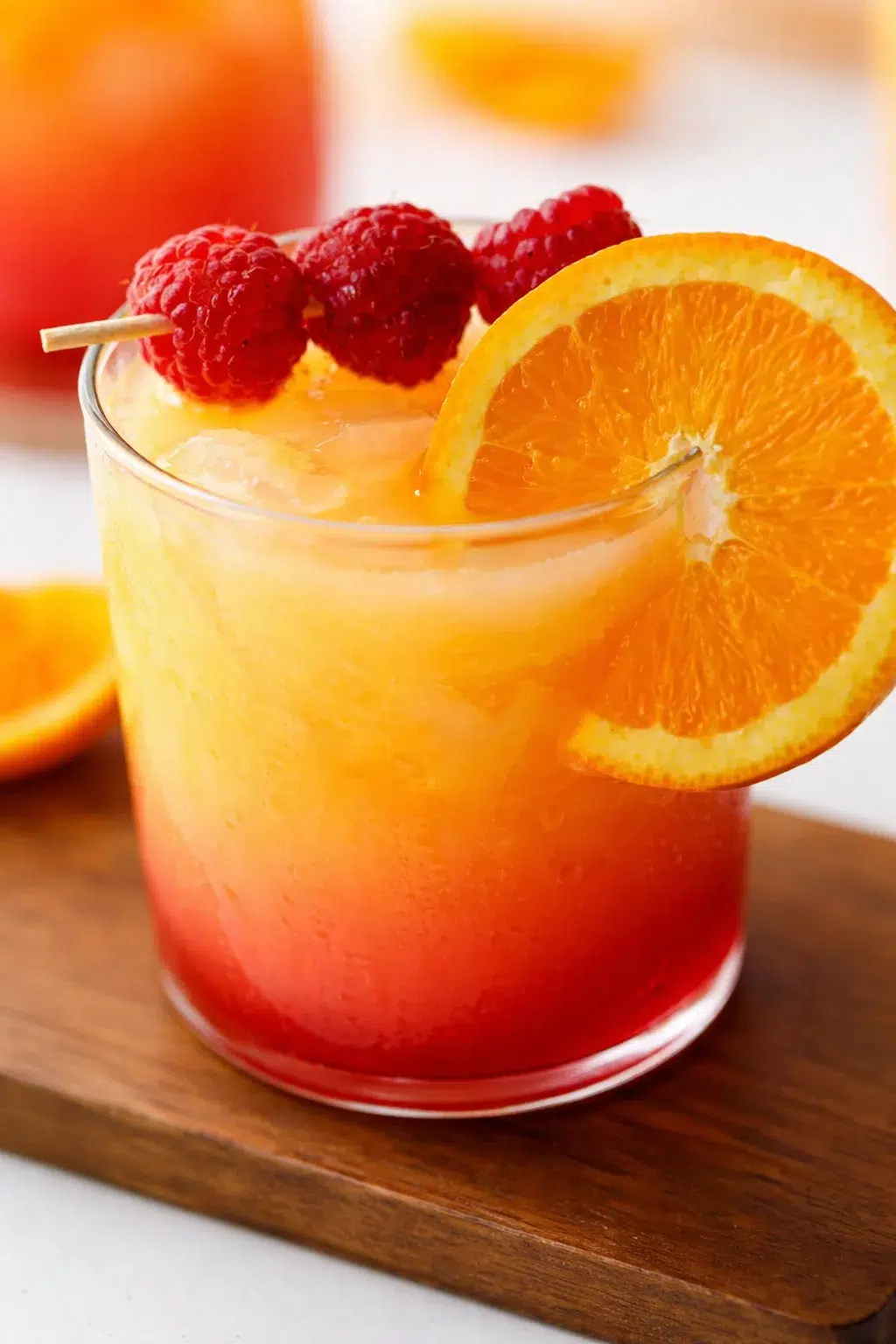 Close-up of a gradient orange-red cocktail with ice, orange slice, and raspberry garnish.