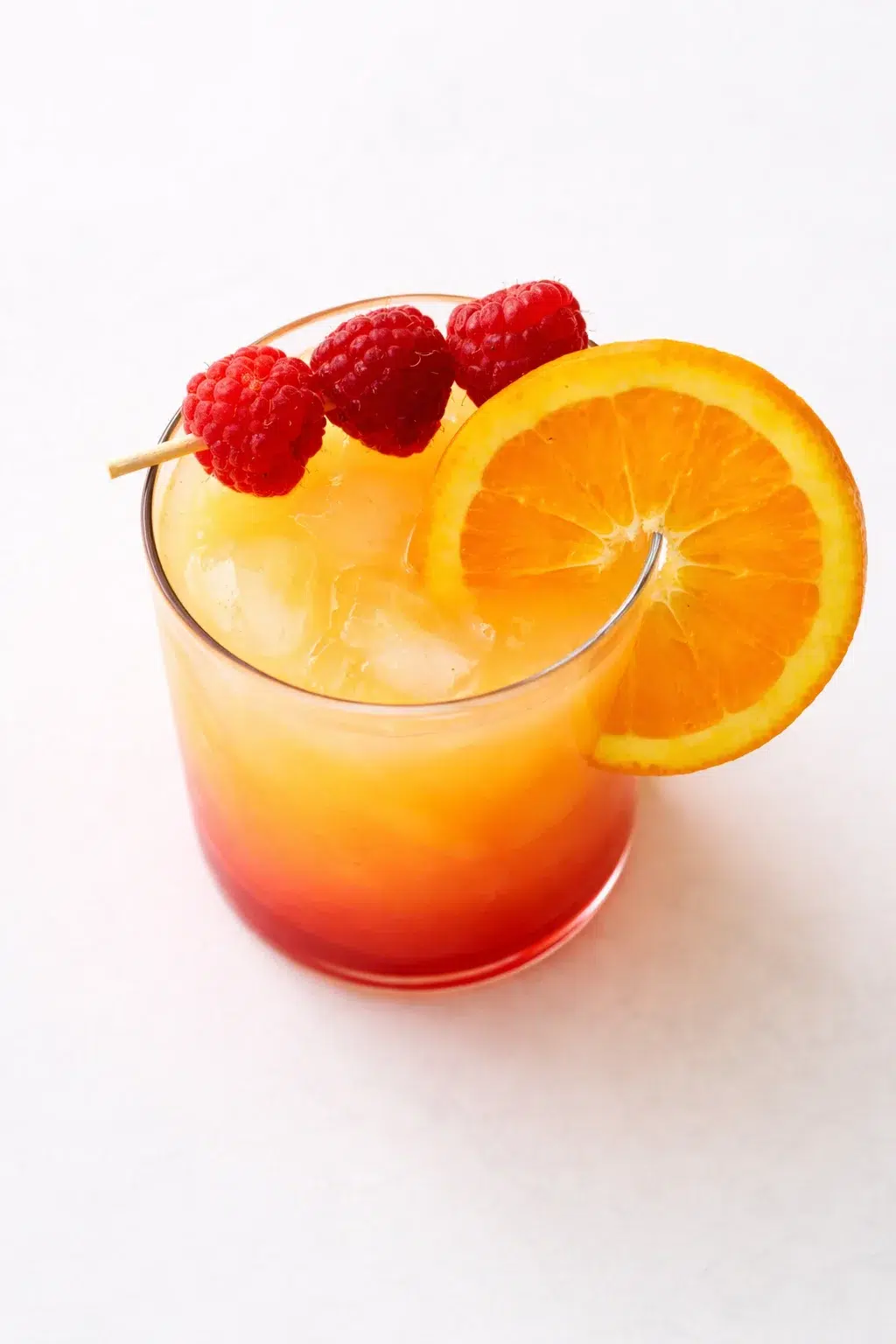 Bright orange-red cocktail in a glass with ice, garnished with an orange slice and raspberry skewers.