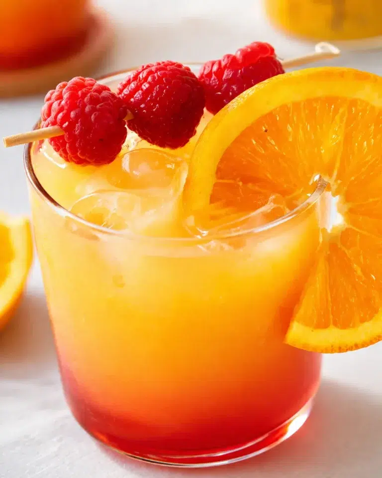 Glass of bright orange sunrise cocktail with ice, orange slice on rim, and raspberry skewered garnish.