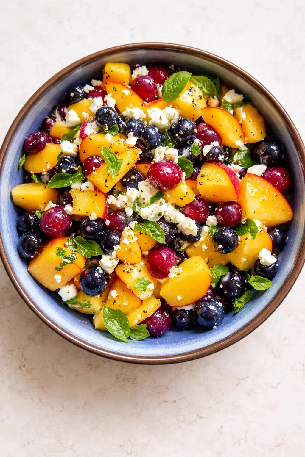 A blue-rimmed bowl filled with peaches, blueberries, grapes, feta, and mint