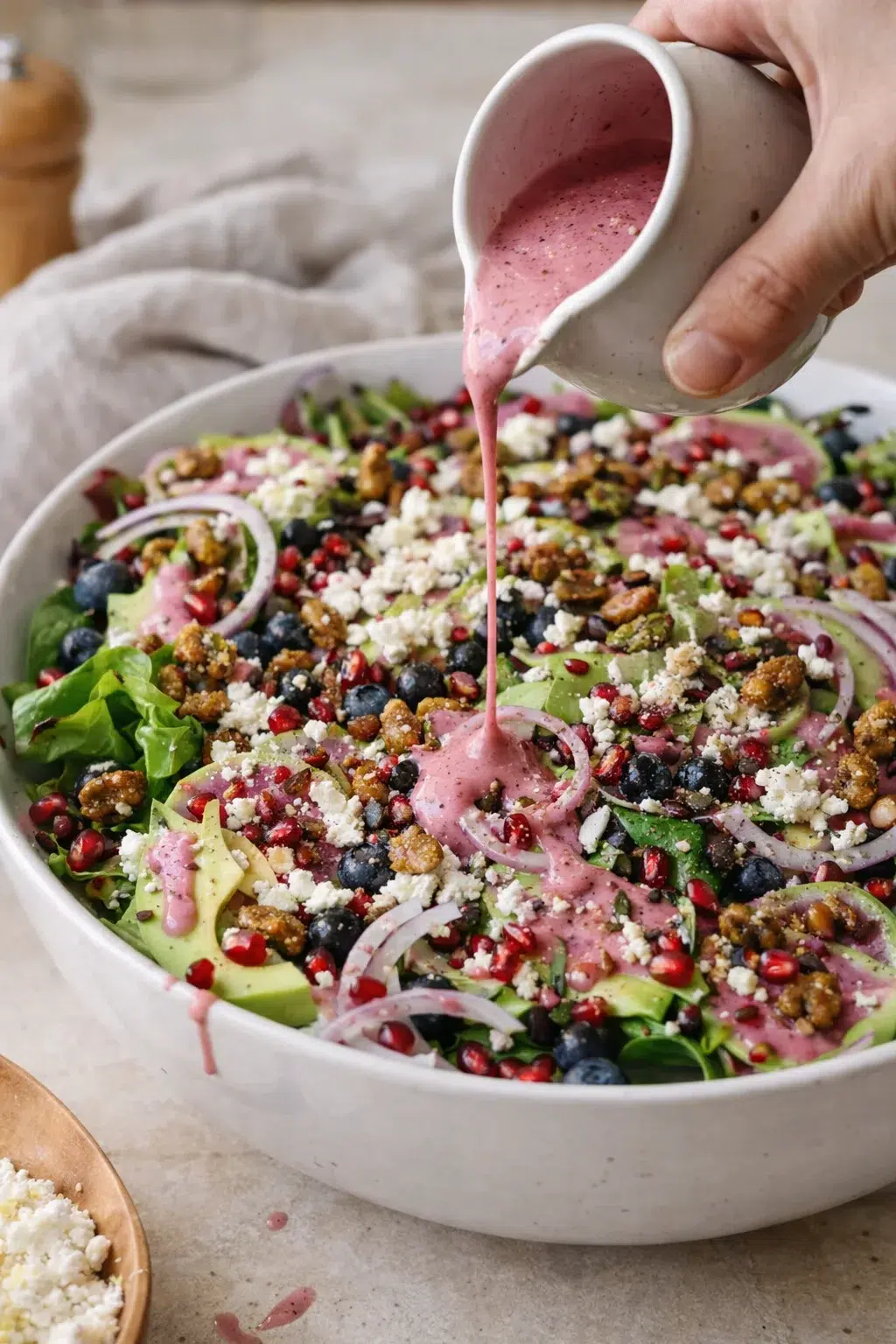 Large white bowl of colorful salad with pink dressing poured from a speckled ceramic pitcher.