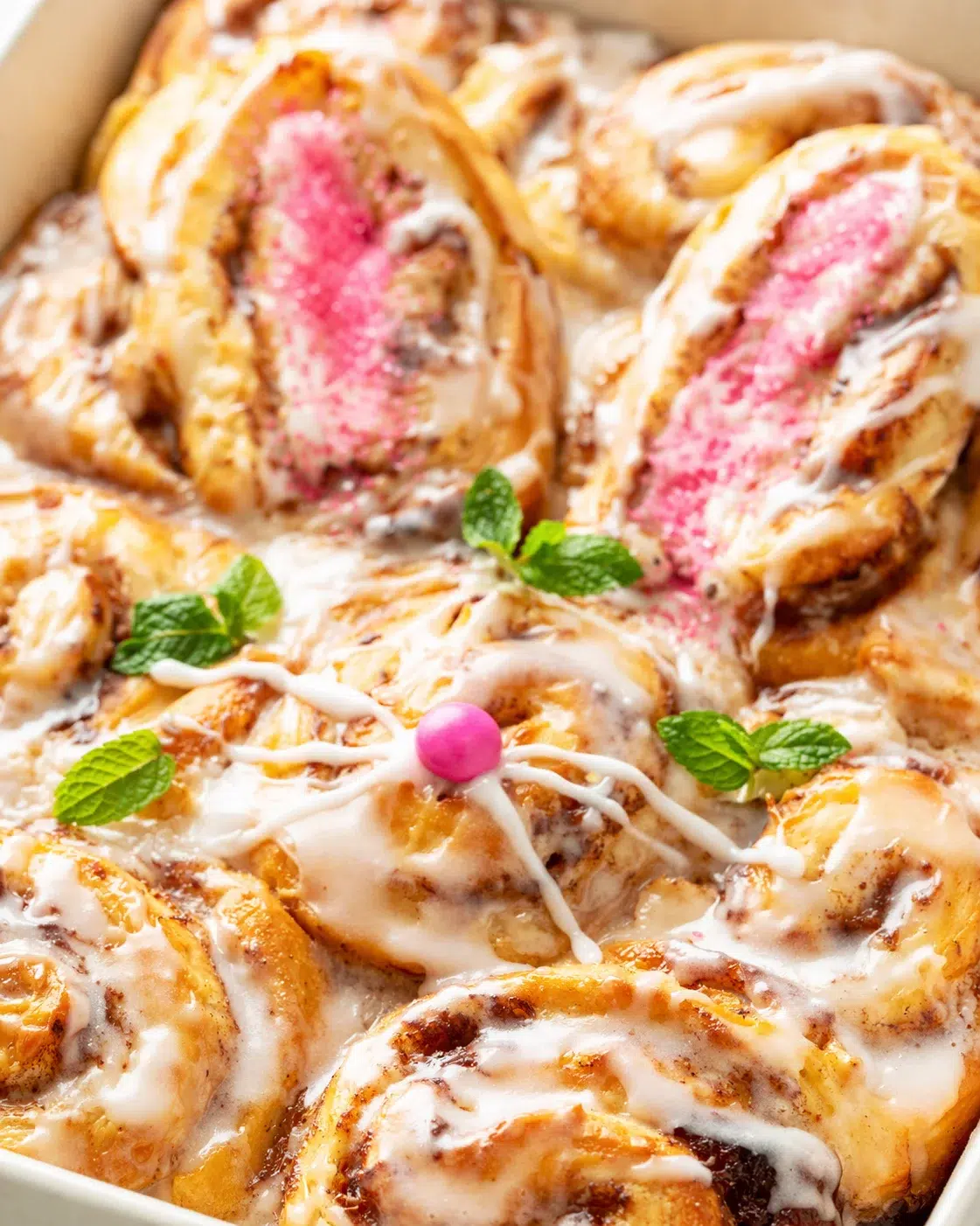 Cinnamon rolls with pink centers, white icing, and mint garnish in a baking dish.