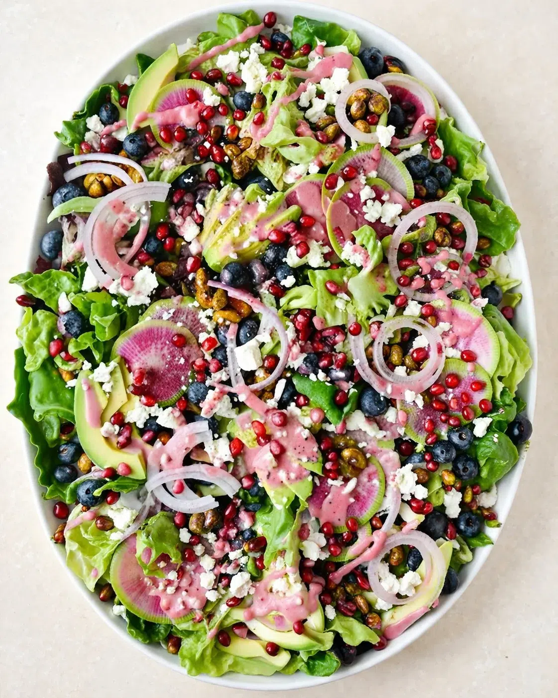 Overhead view of a vibrant green salad with avocado slices, feta, pomegranate seeds, blueberries, onion rings, radish, and pink dressing.