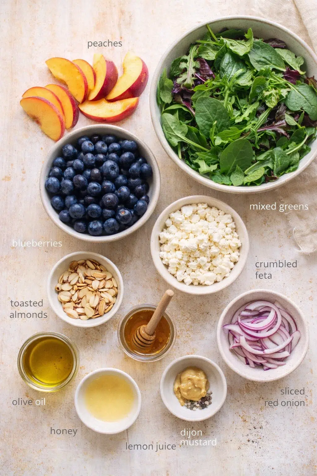 Overhead layout of bowls with peaches, blueberries, greens, feta, almonds, onions, and dressings like olive oil and honey.