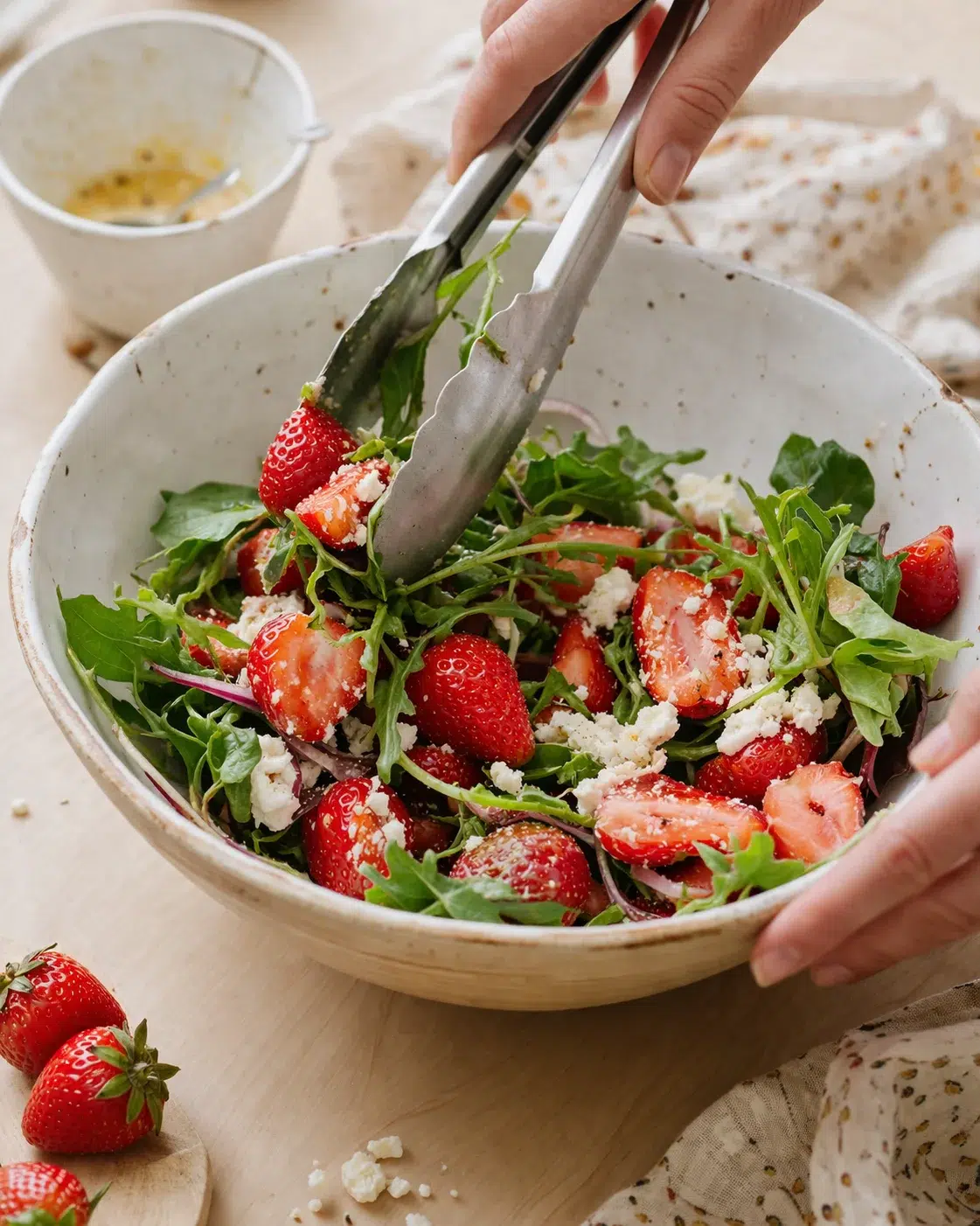 Rustic ceramic bowl of strawberry arugula salad with feta, being tossed with tongs.