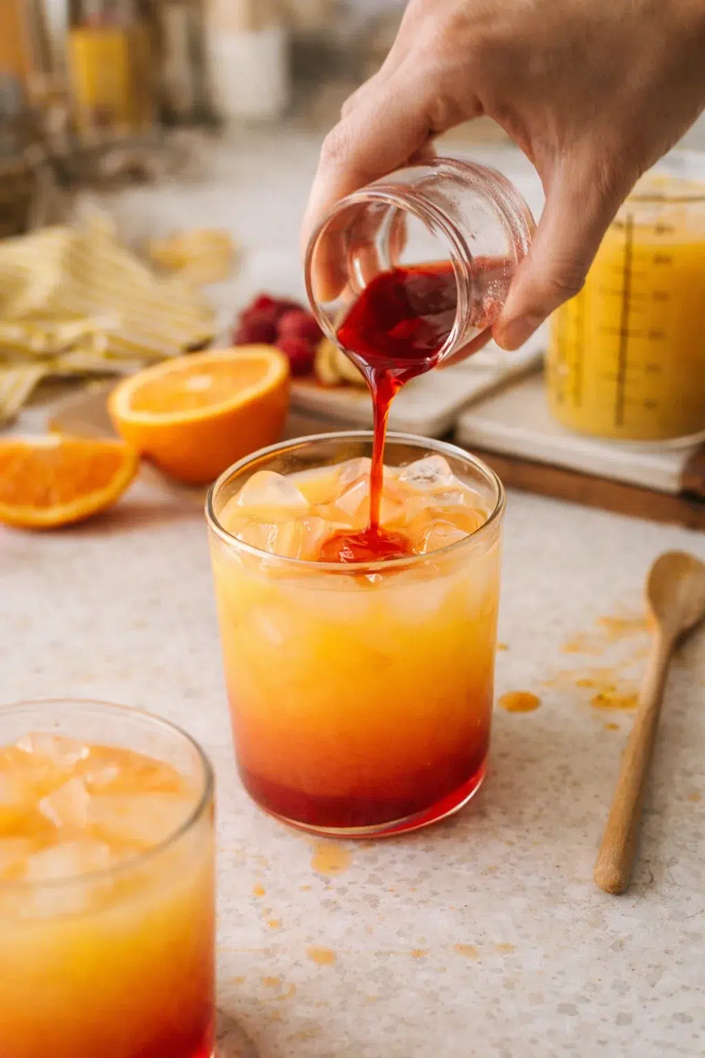 hand pouring red syrup into orange iced drink on countertop