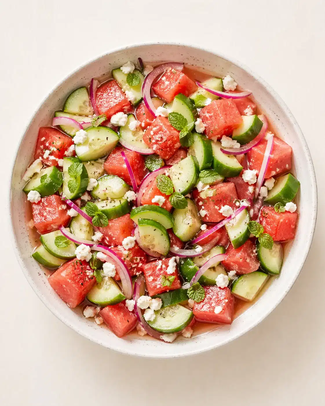 Overhead view of a watermelon-cucumber-feta salad in a speckled white bowl.