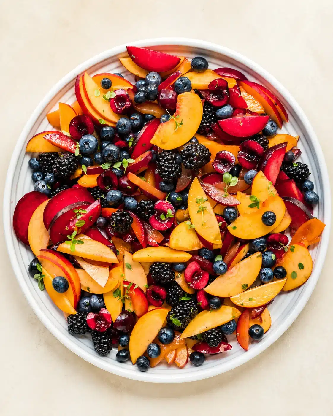 top-down view of a white bowl filled with sliced peaches, plums, blueberries, blackberries, and microgreens