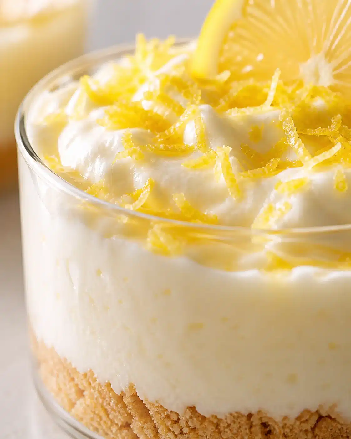 Close-up of lemon cheesecake in a glass with a crumb crust, creamy filling, lemon zest, and a lemon wedge.