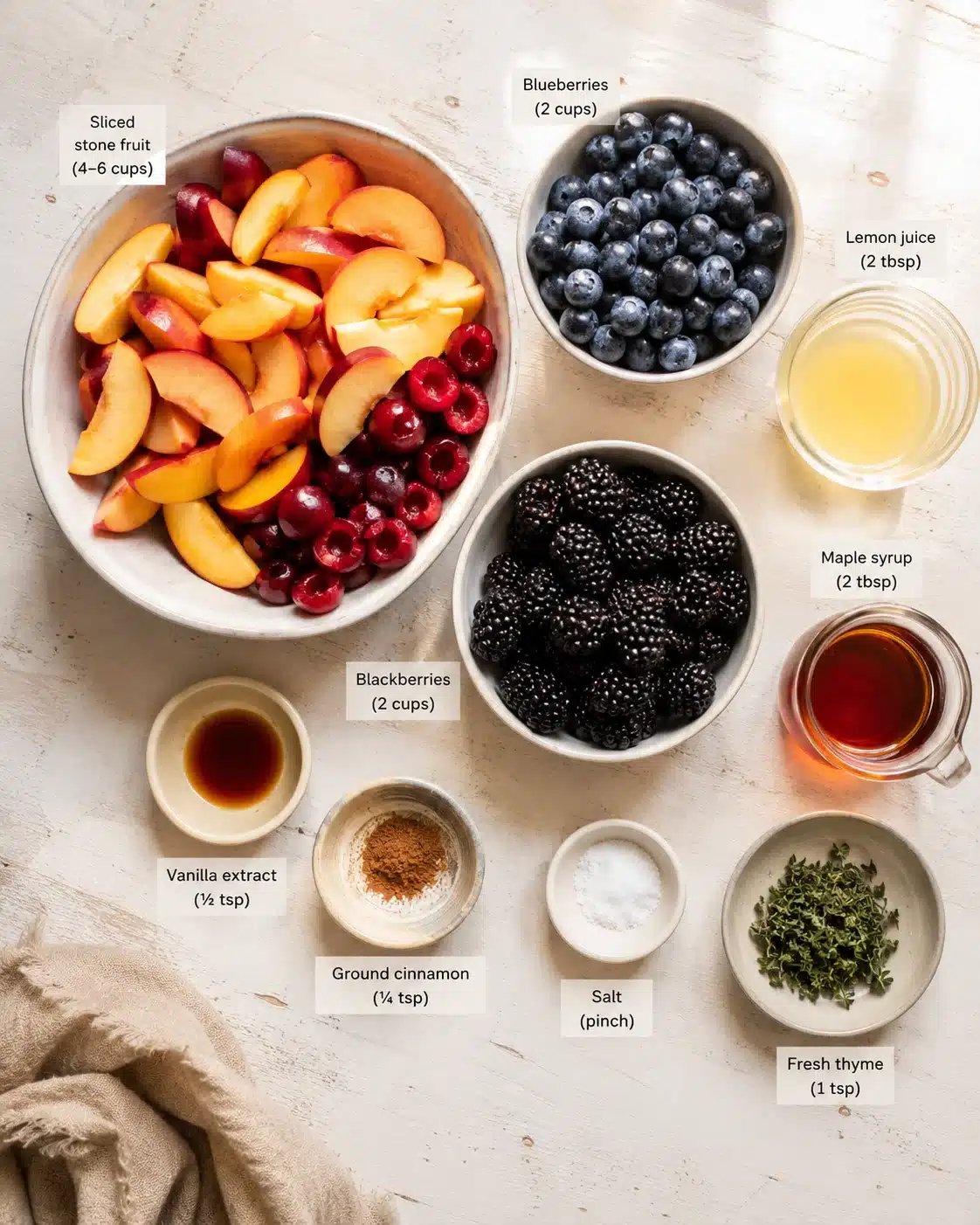 Overhead view of sliced stone fruit, blueberries, and blackberries in bowls with labeled ingredients.