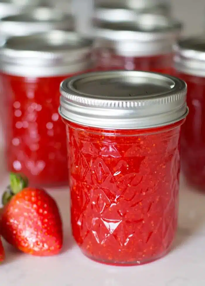 red strawberry jam in embossed mason jars with fresh strawberries nearby on a light surface