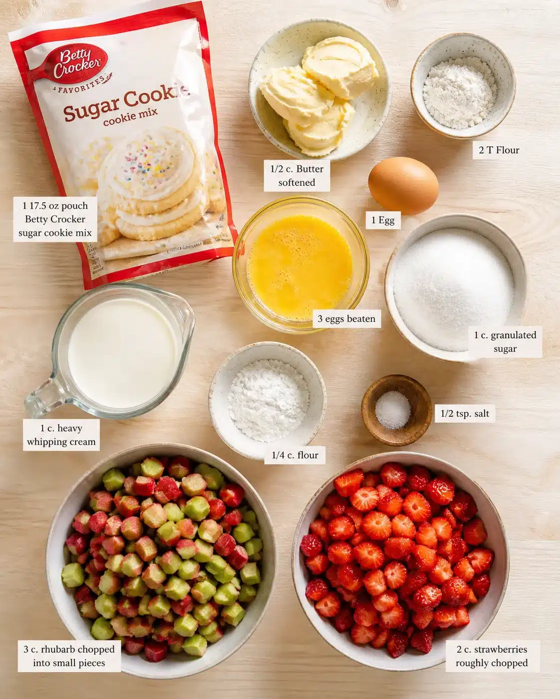 Flat lay of baking ingredients on wooden table: cookie mix bag, butter, eggs, flour, sugar, cream, chopped rhubarb, and strawberries.